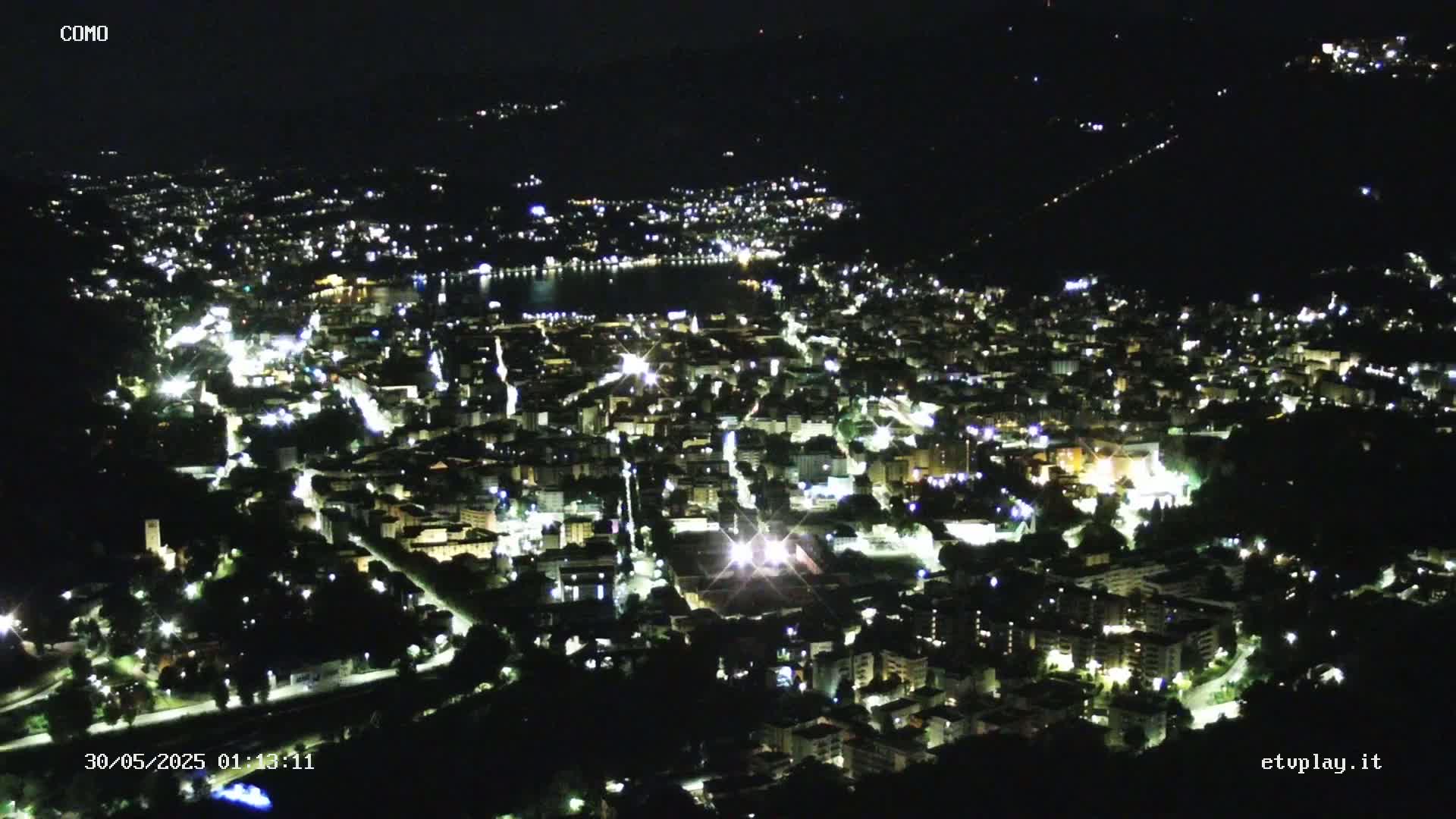 A nighttime aerial view of a city illuminated by streetlights and building lights, nestled beside a dark body of water under a clear, dark sky.