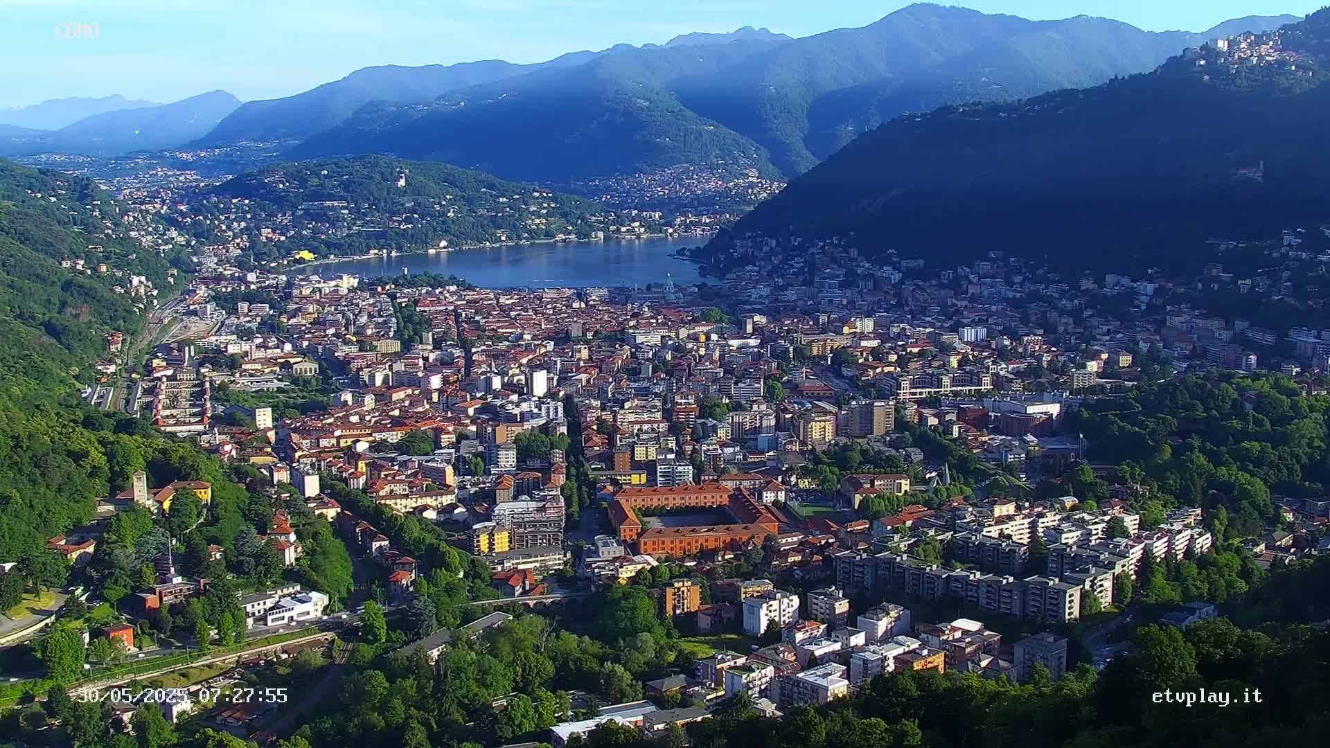 An aerial view shows a city nestled between mountains and a lake under a clear, sunny sky.