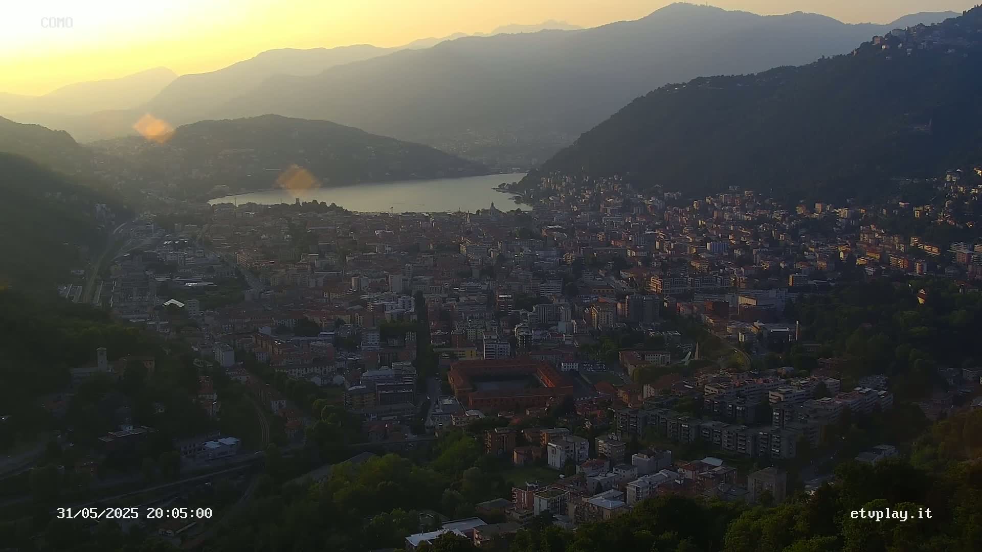 A sprawling city nestled between mountains and a lake at sunset, under a hazy sky.