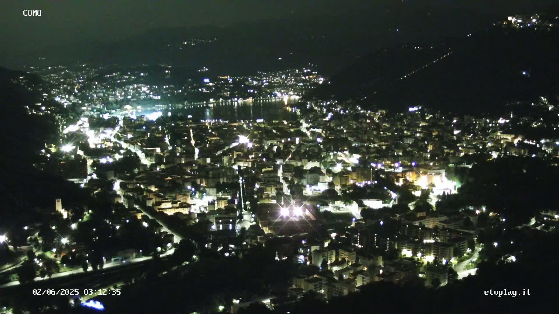 A nighttime aerial view of a city nestled beside a body of water, illuminated by numerous lights.