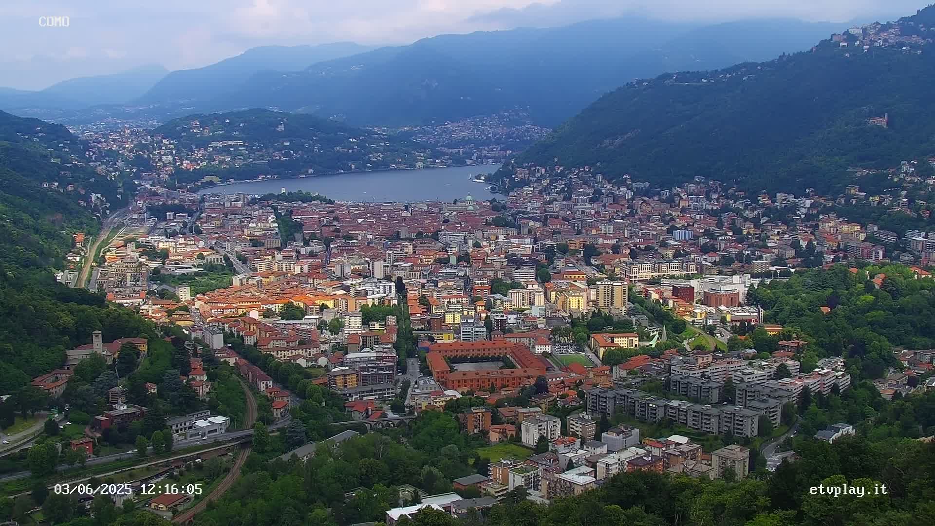 An aerial view shows a city nestled between mountains and a lake under partly cloudy skies.