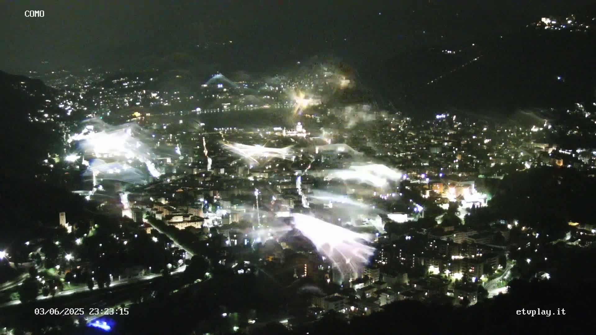 A nighttime aerial view of a city illuminated by numerous bright lights and streaks of white light, possibly fireworks, emanating from various locations.