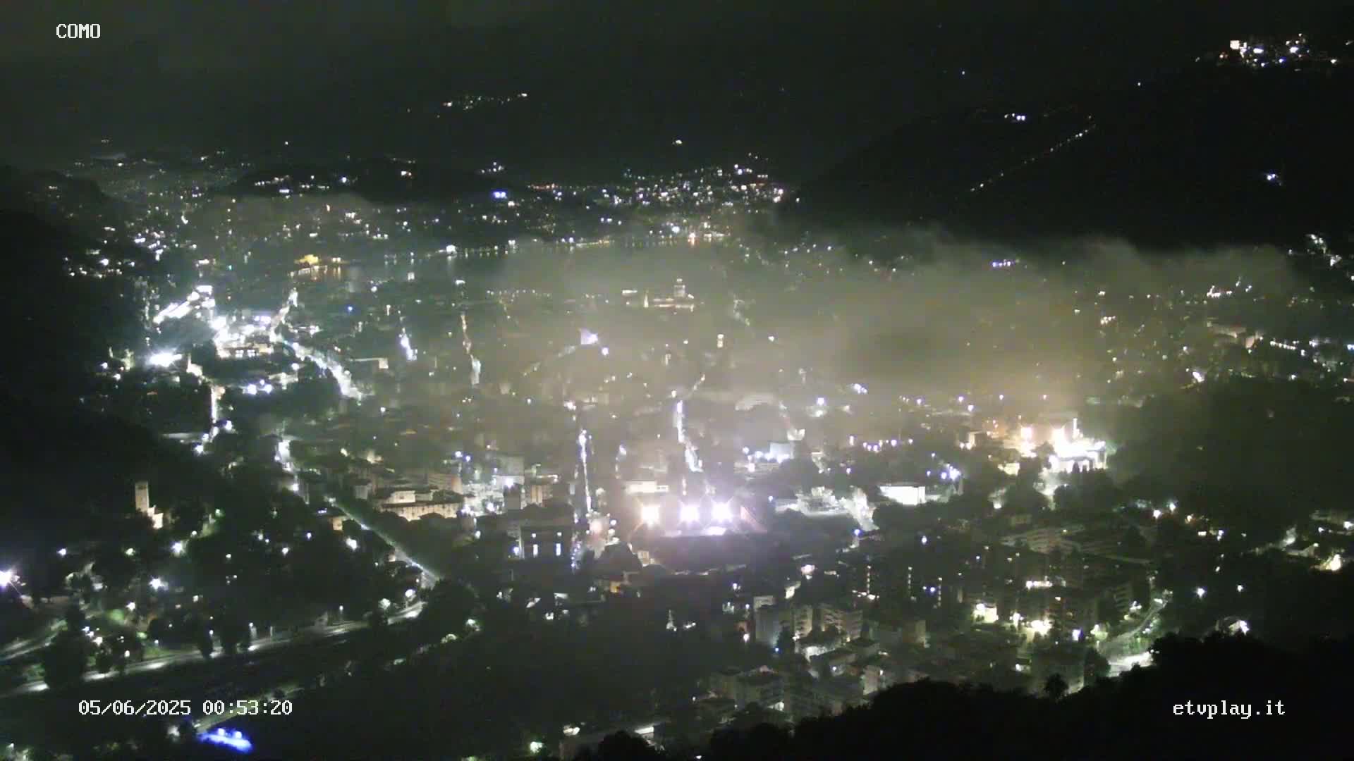 A nighttime, high-angle view of a city valley shrouded in fog, with numerous lights illuminating the buildings and streets below.