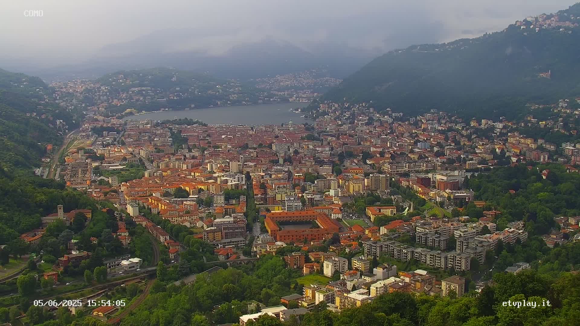 An aerial view shows a city nestled between a lake and mountains on an overcast day.