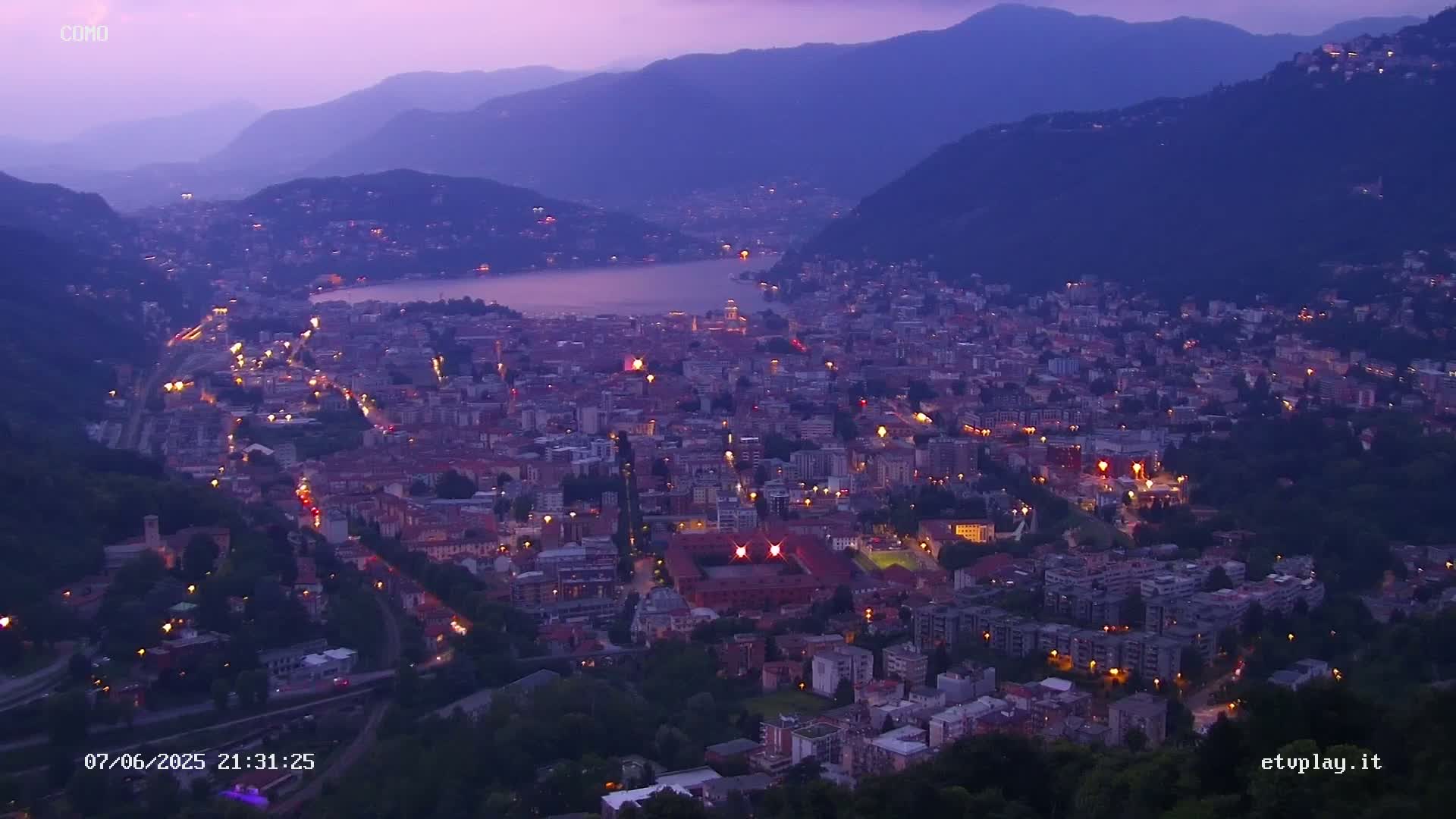 A nighttime view of a city nestled between mountains and a lake under a dusky purple sky.
