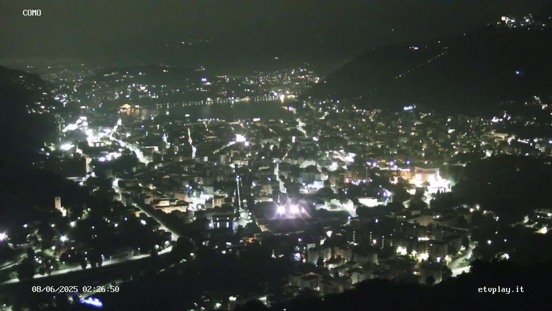 A nighttime, high-angle view of a city illuminated by numerous lights, situated beside a dark body of water, under a clear sky.