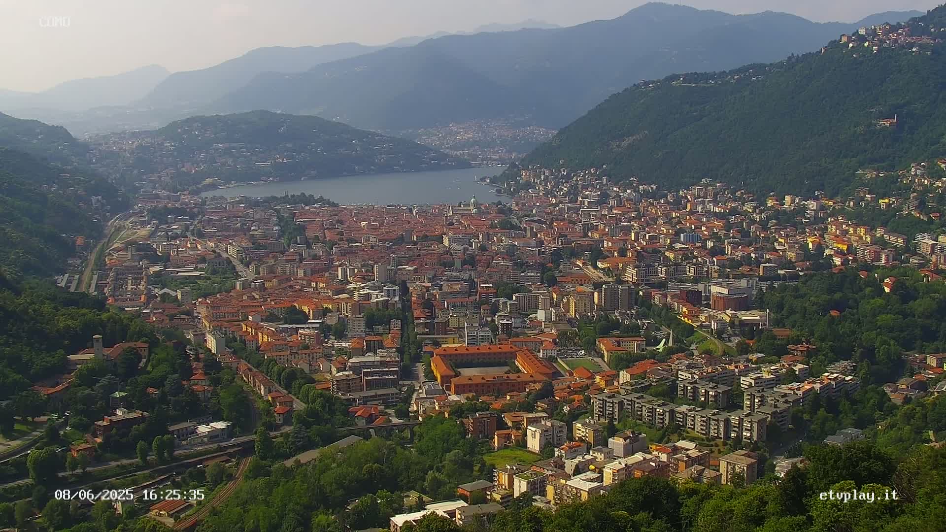 A hazy, sunny day reveals a sprawling city nestled in a valley between mountains, next to a calm lake.