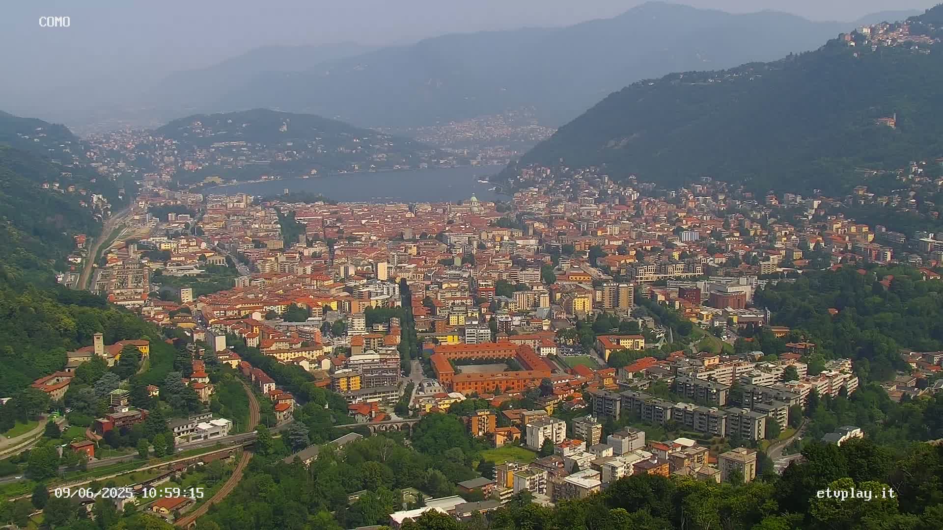A hazy, sunny day reveals a sprawling city nestled in a valley beside a lake, surrounded by lush green hills.