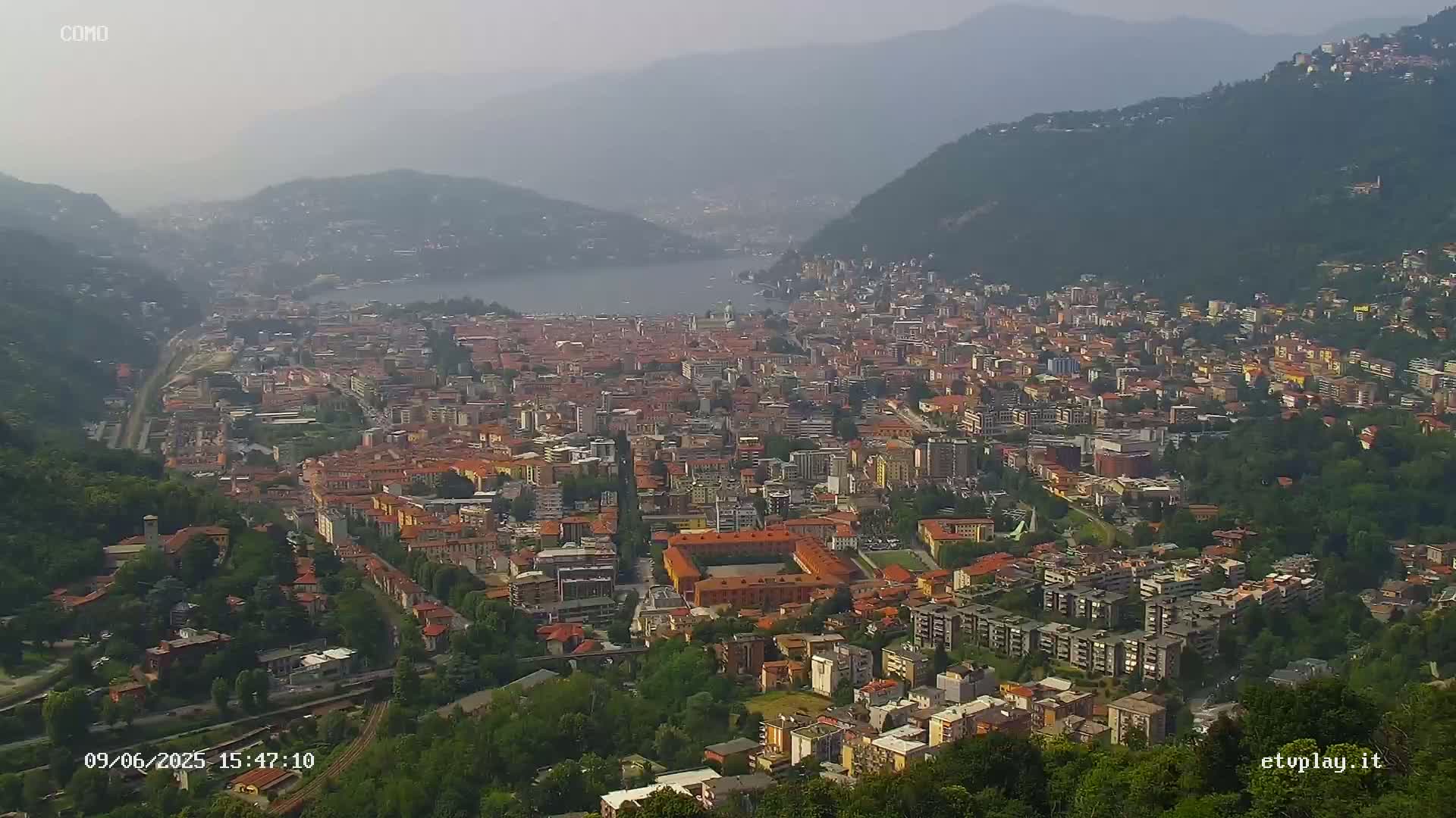 An aerial view shows a hazy city nestled between hills and a lake under a hazy sky.