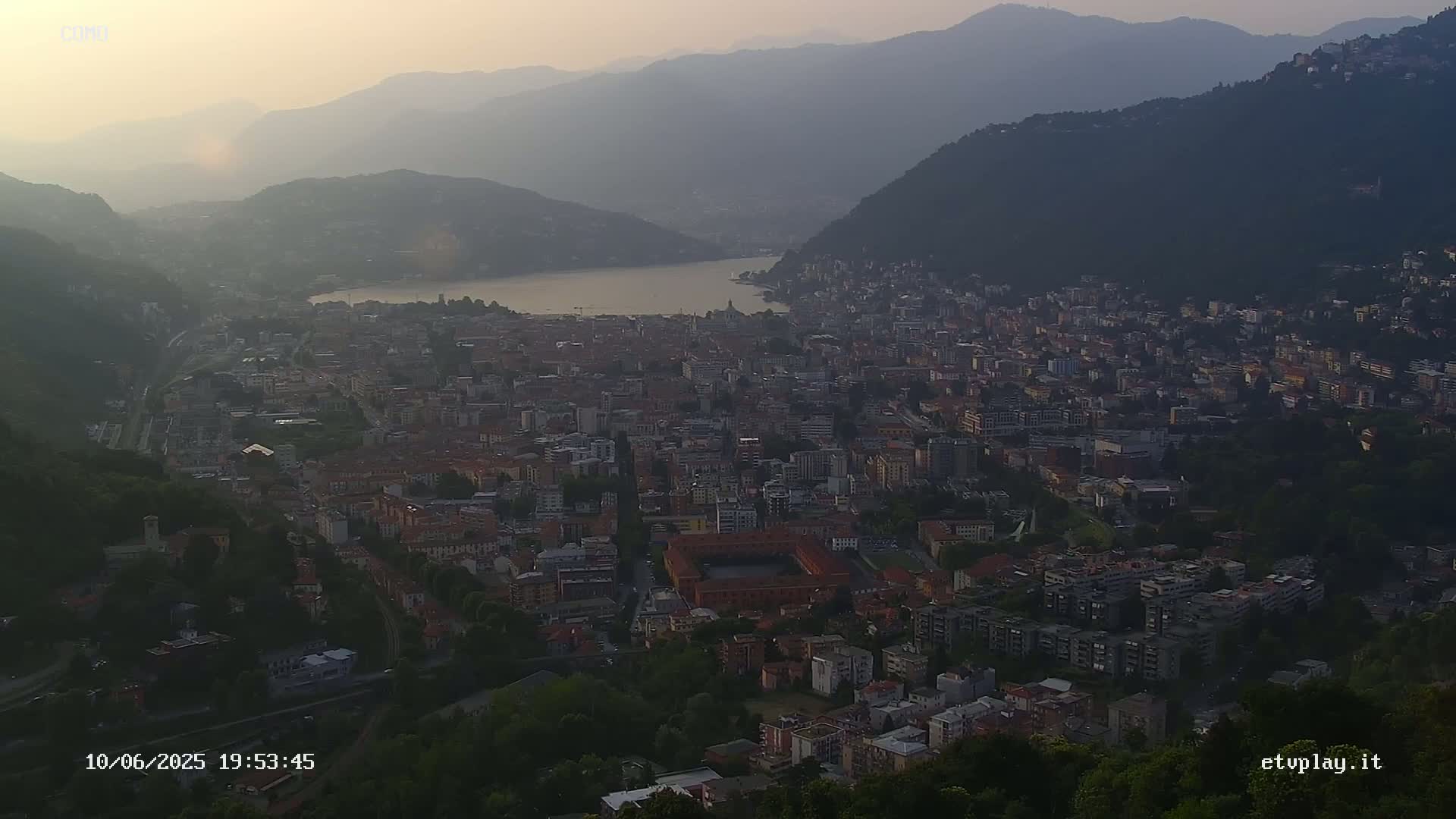 A hazy sunset view overlooks a sprawling city nestled between mountains and a lake.