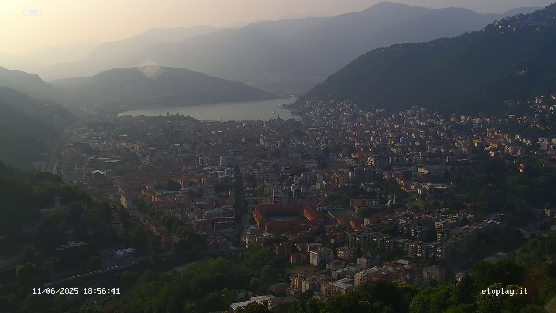 A hazy sunset view overlooks a city nestled between hills and a lake.