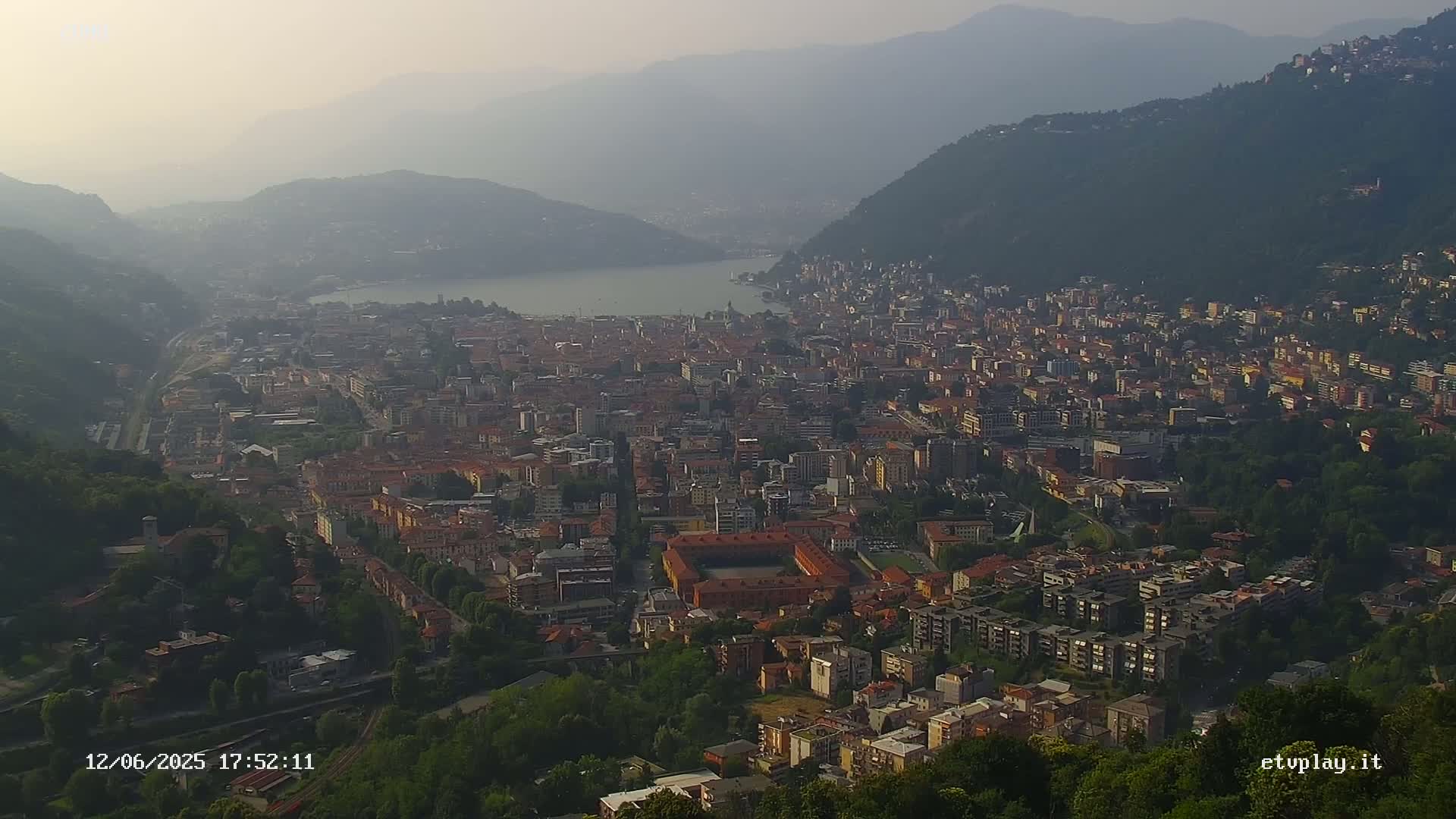 A hazy view of a city nestled between mountains and a lake, with green hillsides surrounding the urban area.