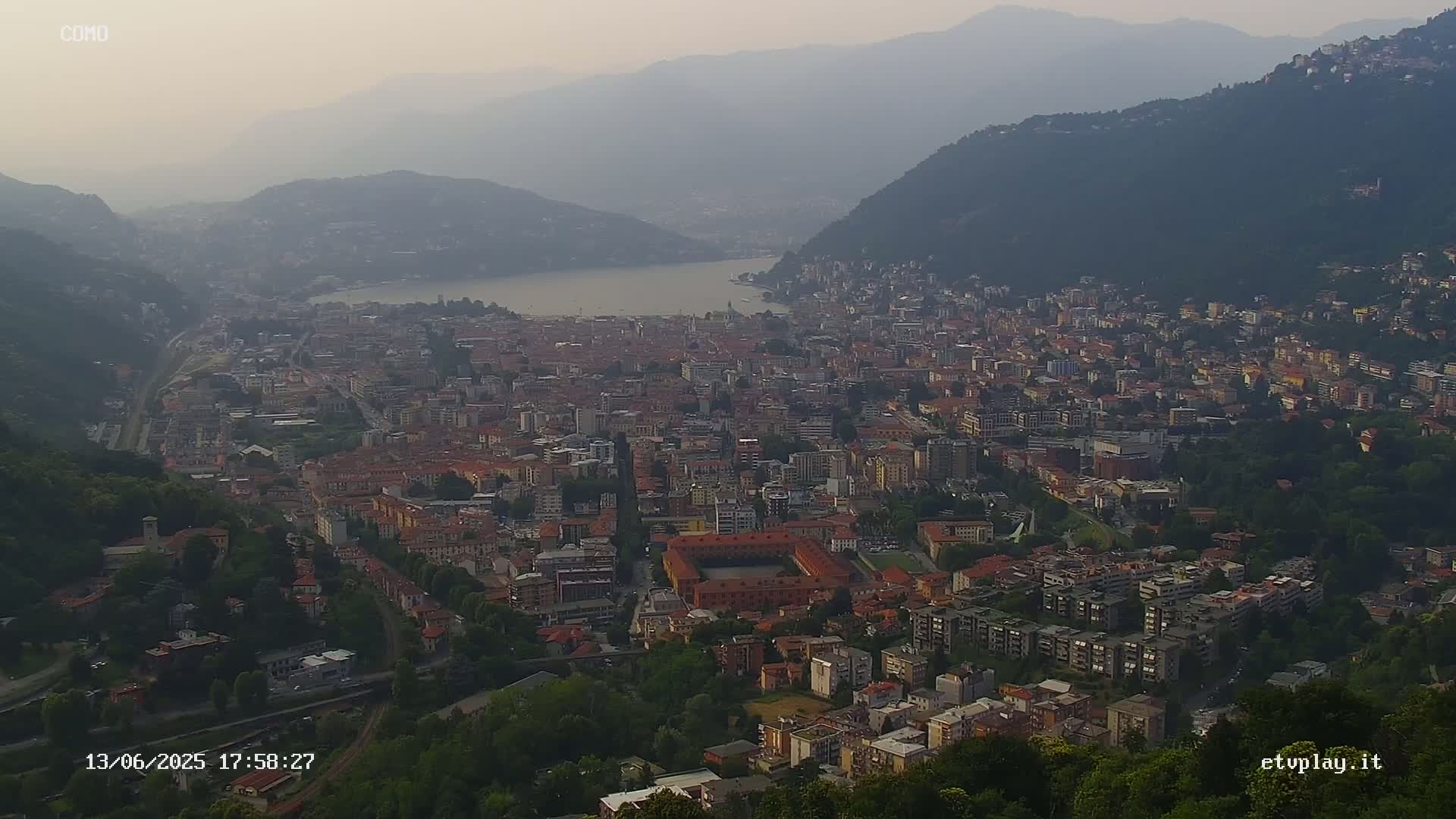 A hazy view from a hilltop overlooking a large city nestled between mountains and a lake.