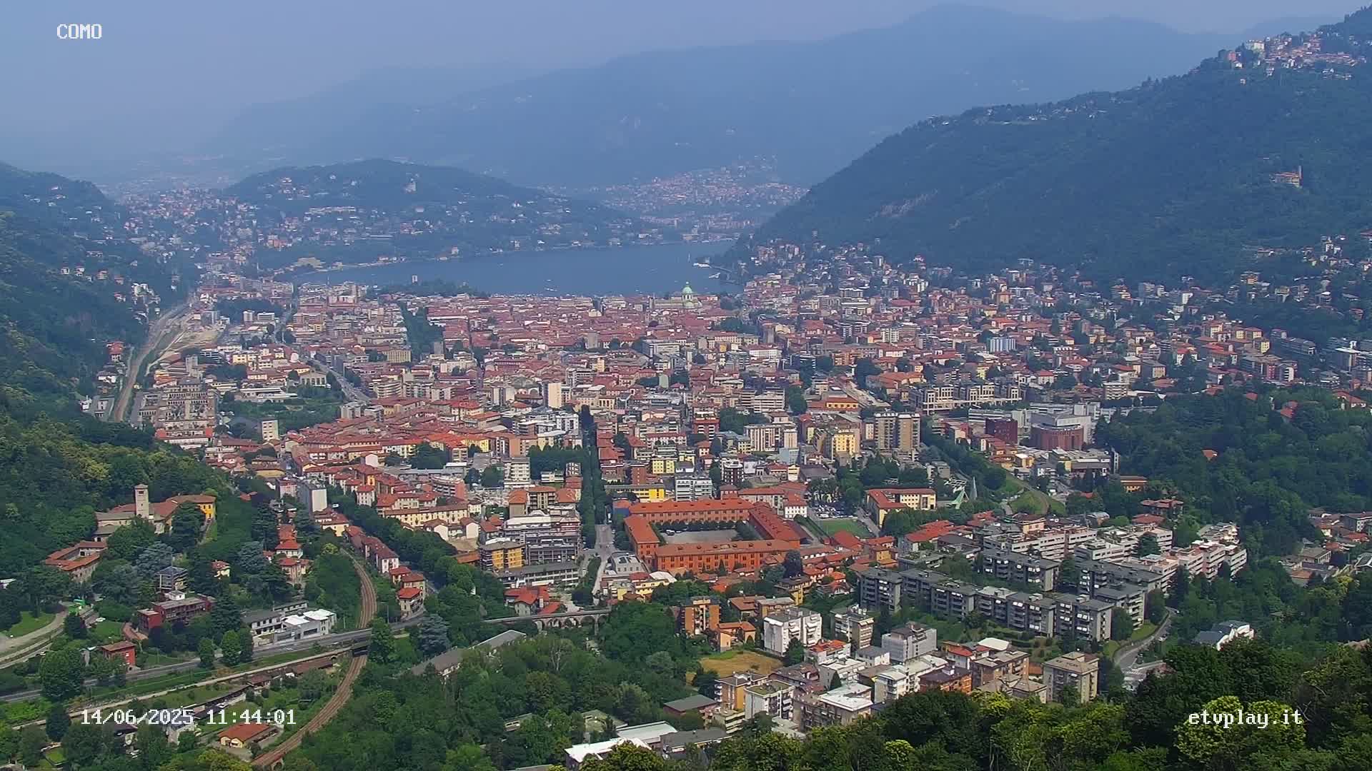 A sprawling city nestled in a valley beside a lake, viewed from a high vantage point on a hazy day.