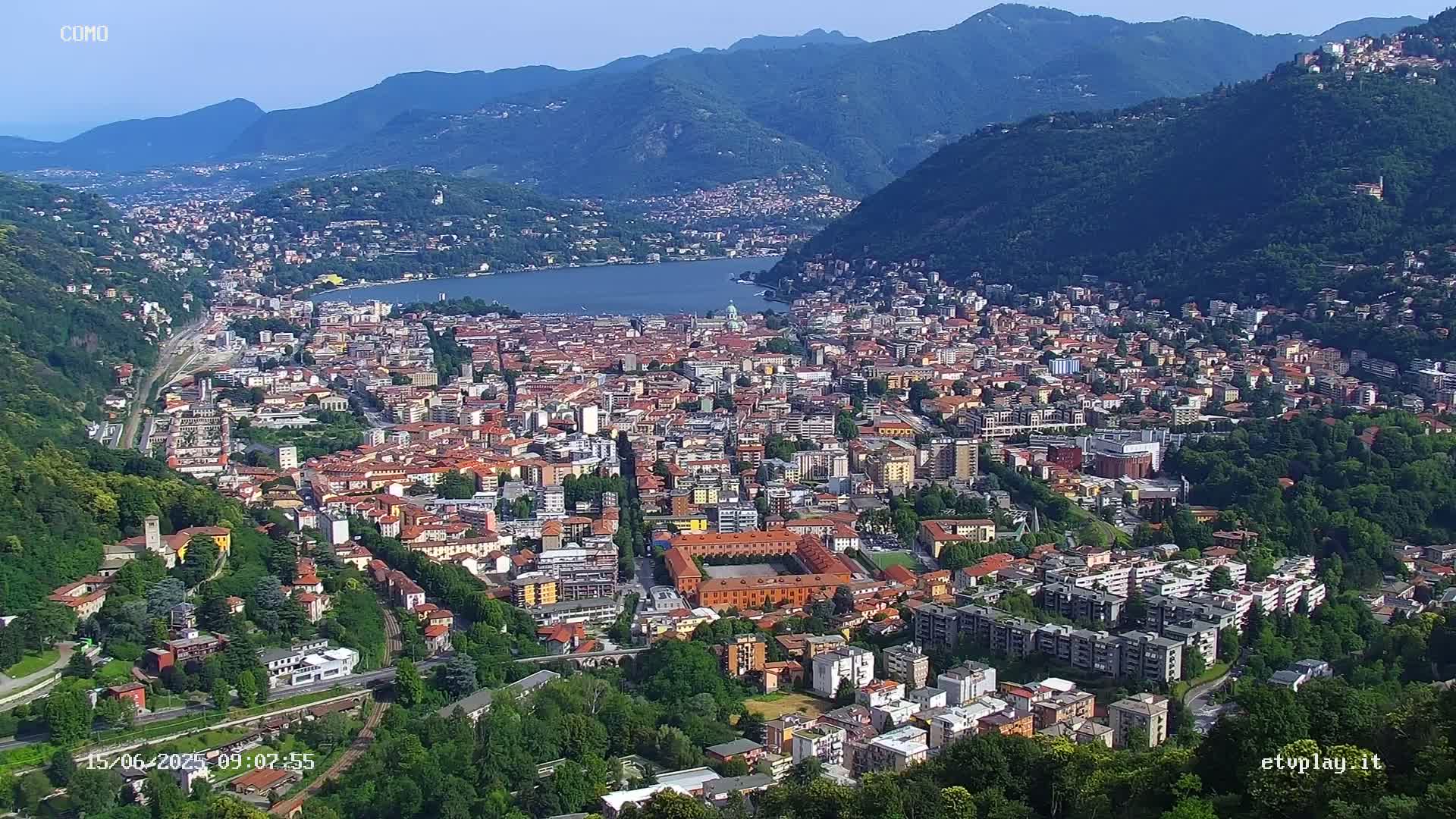 An aerial view of a city nestled in a valley between mountains on a clear, sunny day, with a lake visible in the distance.