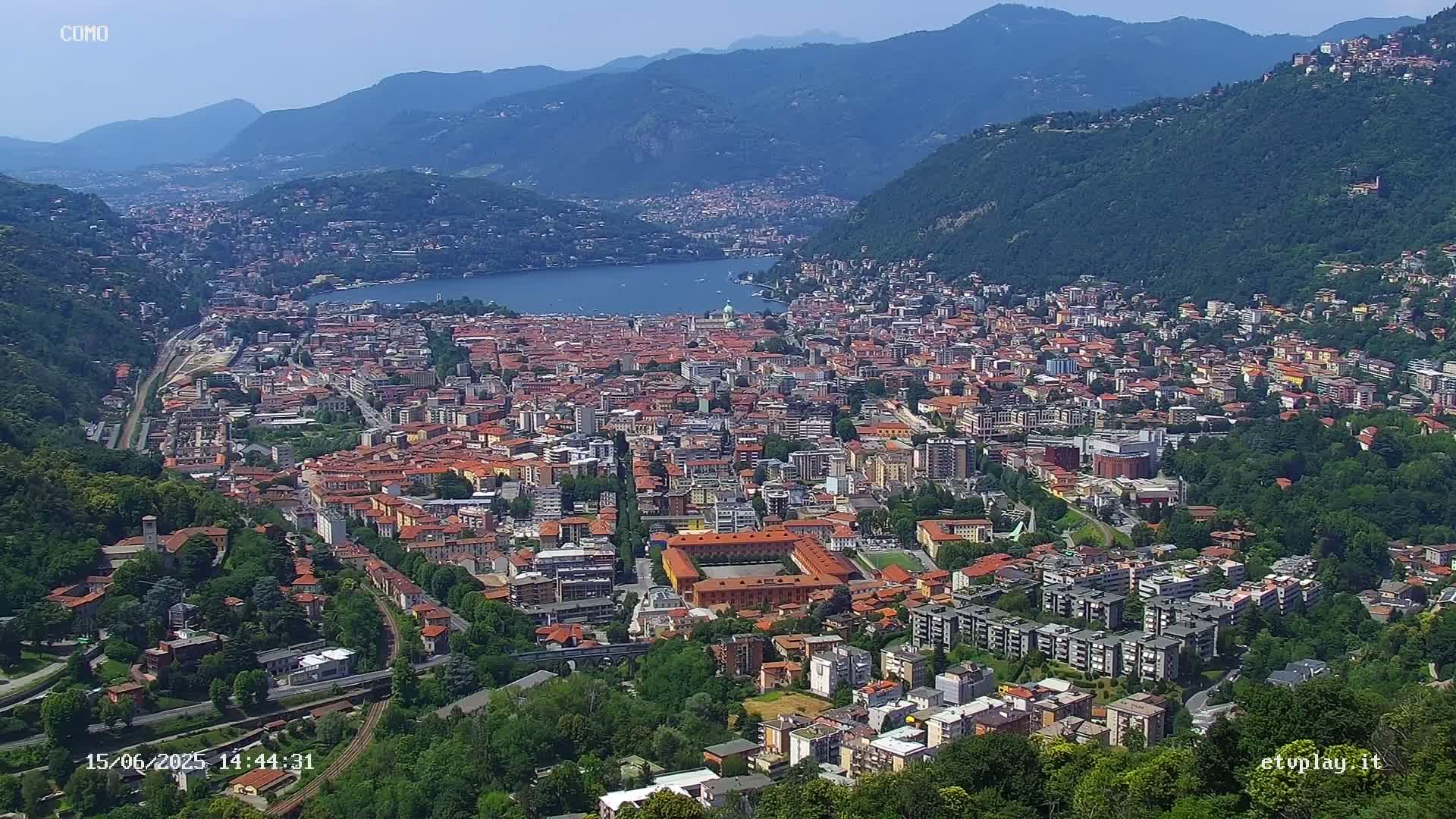 An aerial view of a large city nestled in a valley between mountains on a clear, sunny day, with a lake visible in the distance.