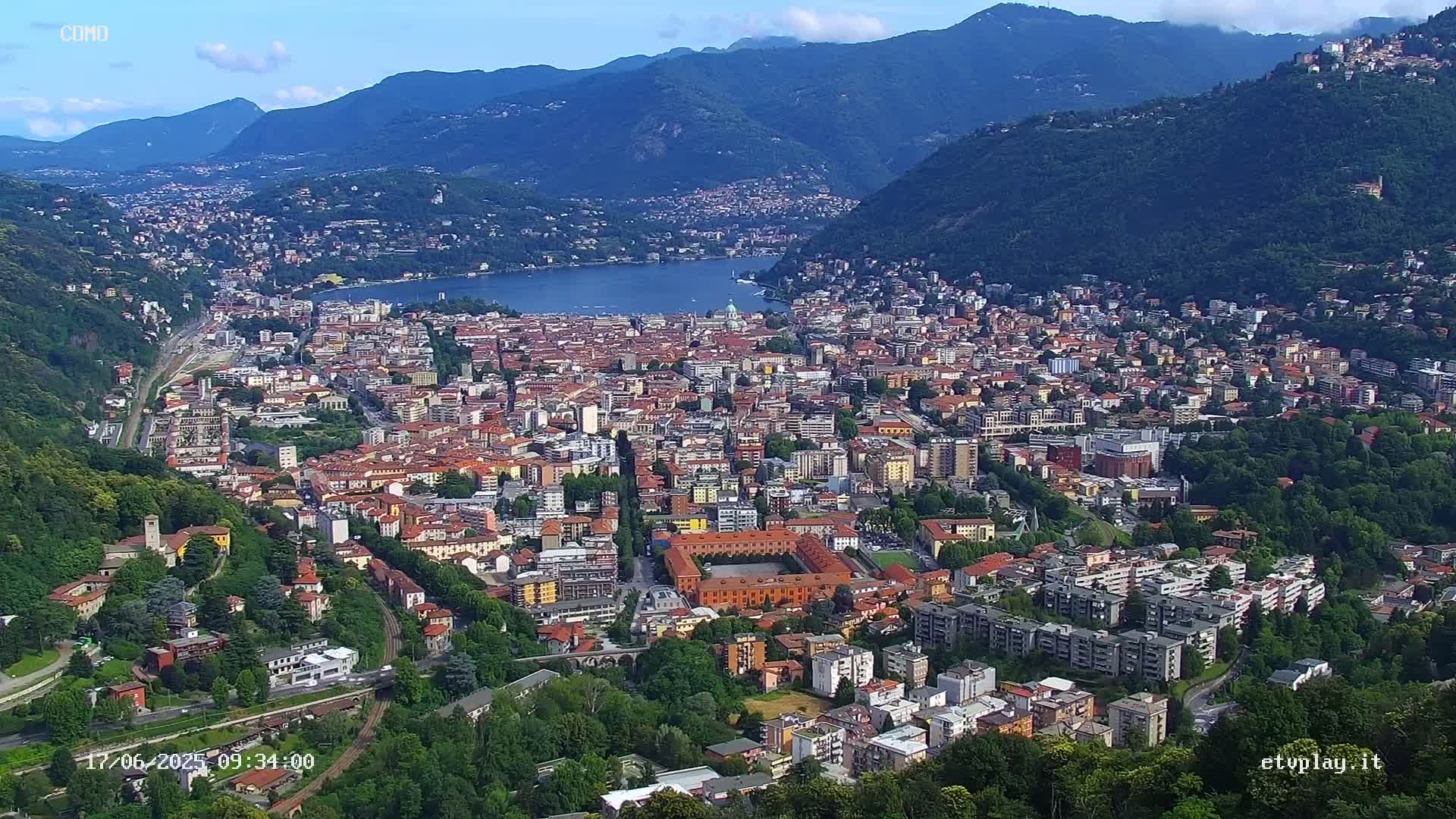 An aerial view shows a city nestled in a valley between mountains, alongside a lake on a sunny day.