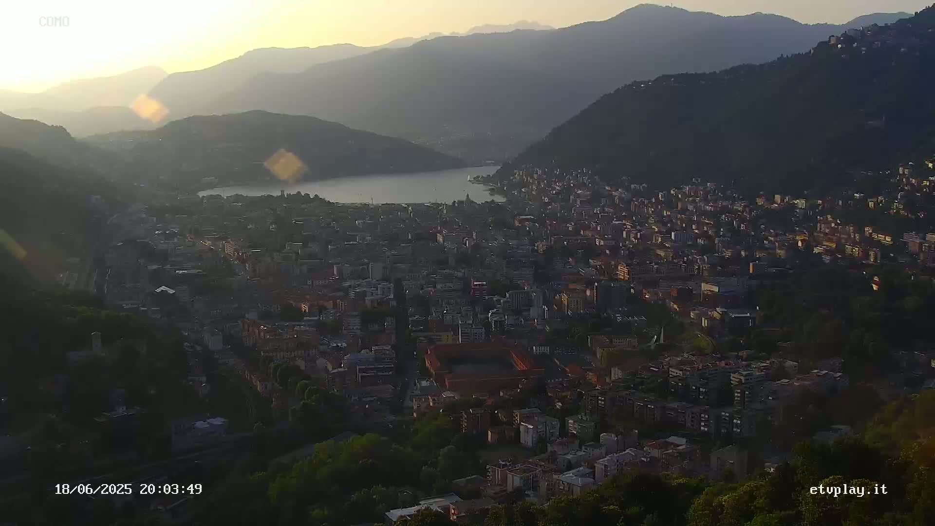 A sun-drenched vista overlooks a city nestled beside a lake and surrounded by mountains.