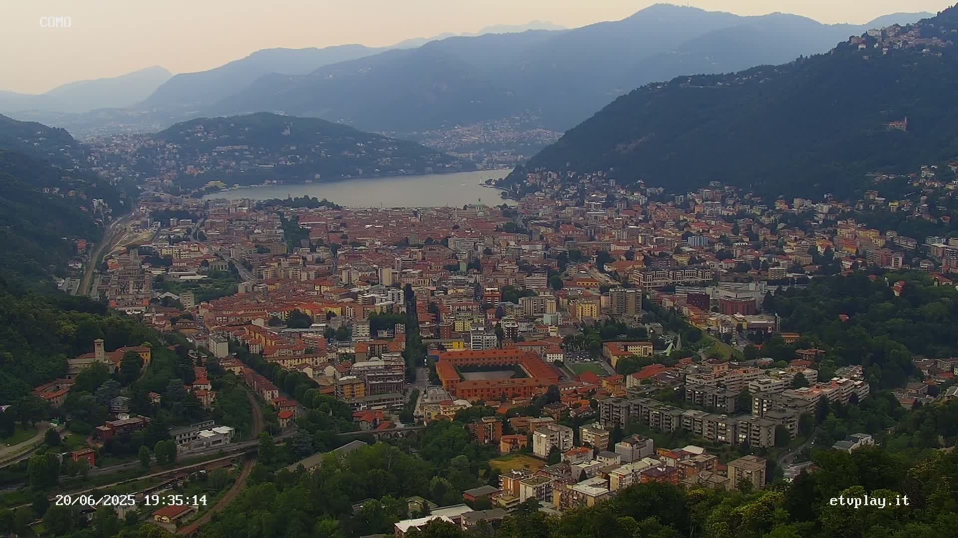 An aerial view of a city nestled between mountains and a lake under a hazy sky.