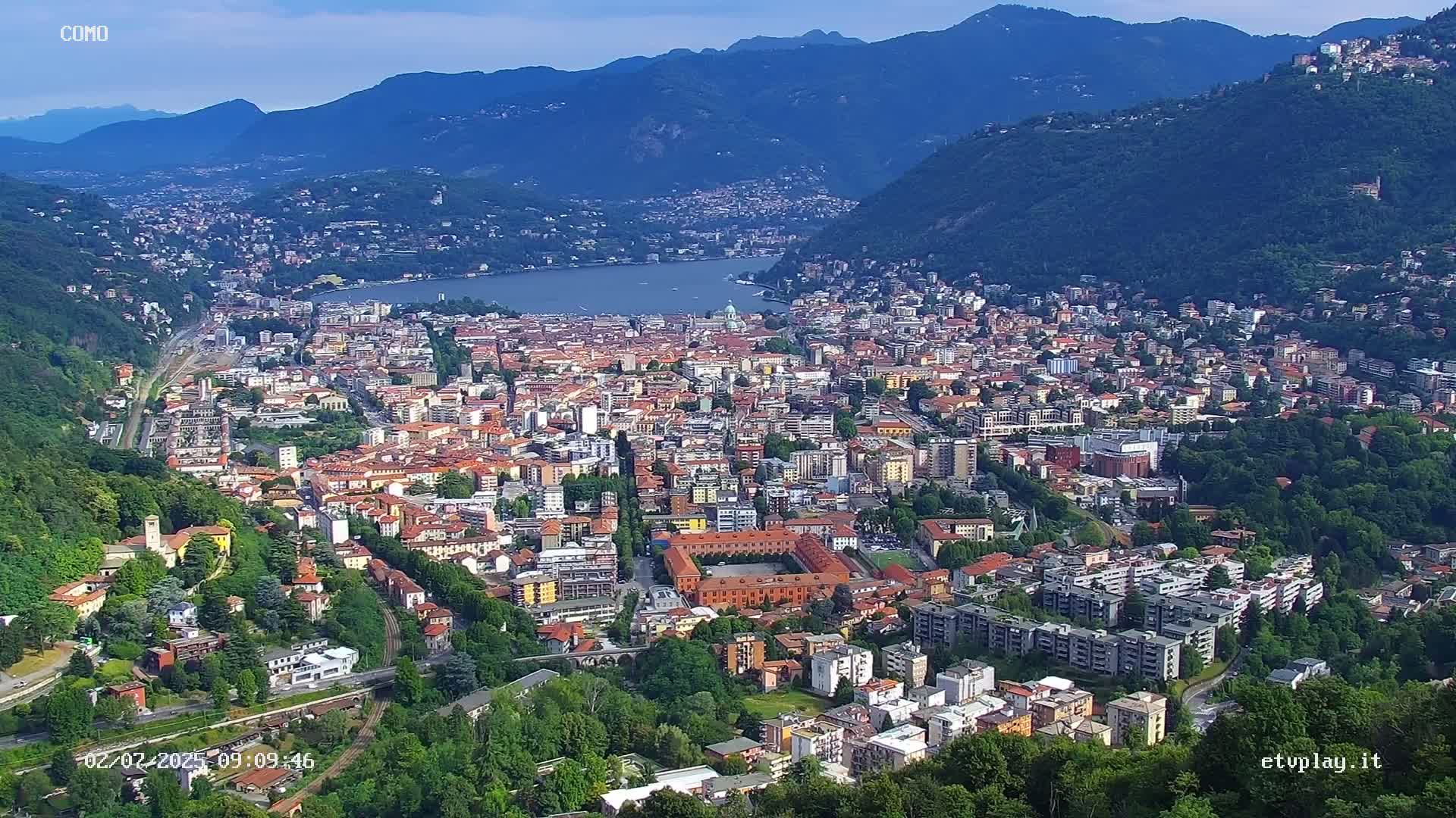 An aerial view shows a city nestled in a valley between mountains on a clear, sunny day, with a lake visible in the distance.