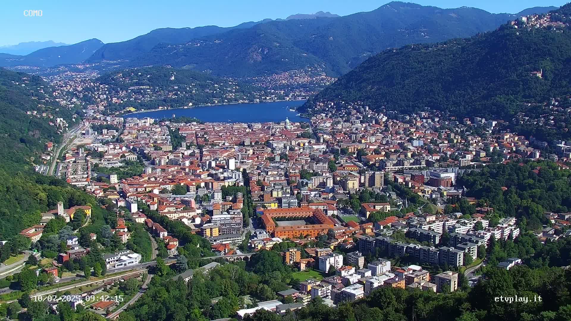 A sprawling city nestled in a valley between mountains, overlooking a calm lake under a clear blue sky.