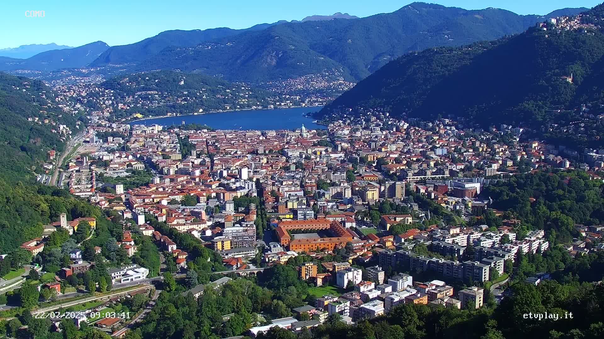 A sprawling city nestled in a valley between mountains, overlooks a lake under sunny skies.
