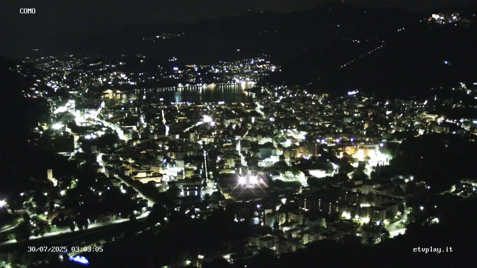 A sprawling city nestled in a valley between mountains, overlooks a lake under sunny skies.