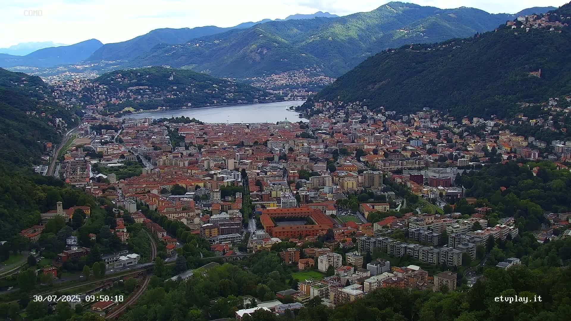 An expansive city nestled in a valley between mountains, overlooks a calm lake under a mostly cloudy sky.