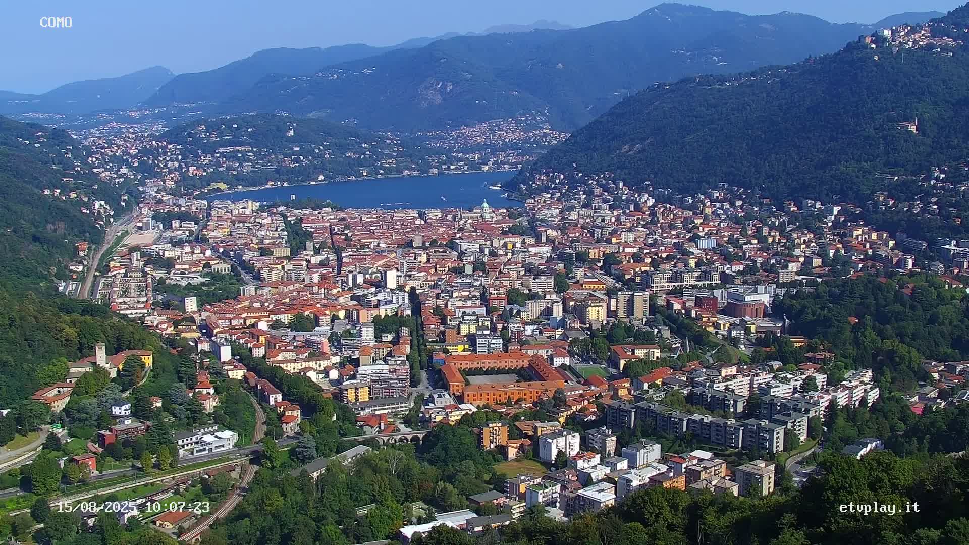 An aerial view shows a sprawling city nestled between mountains and a lake under sunny skies.