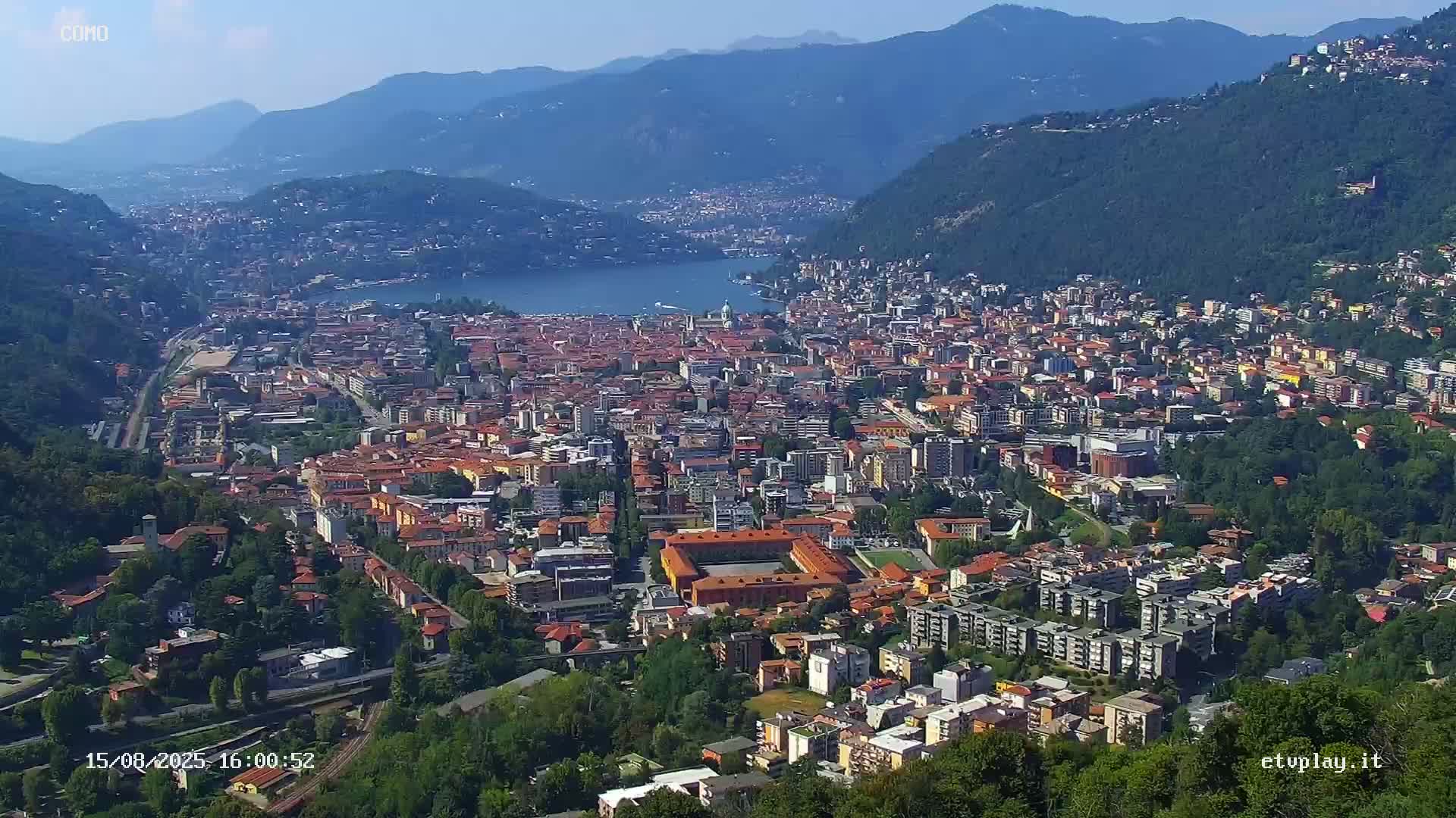 An aerial view shows a sprawling city nestled between mountains and a lake under sunny skies.