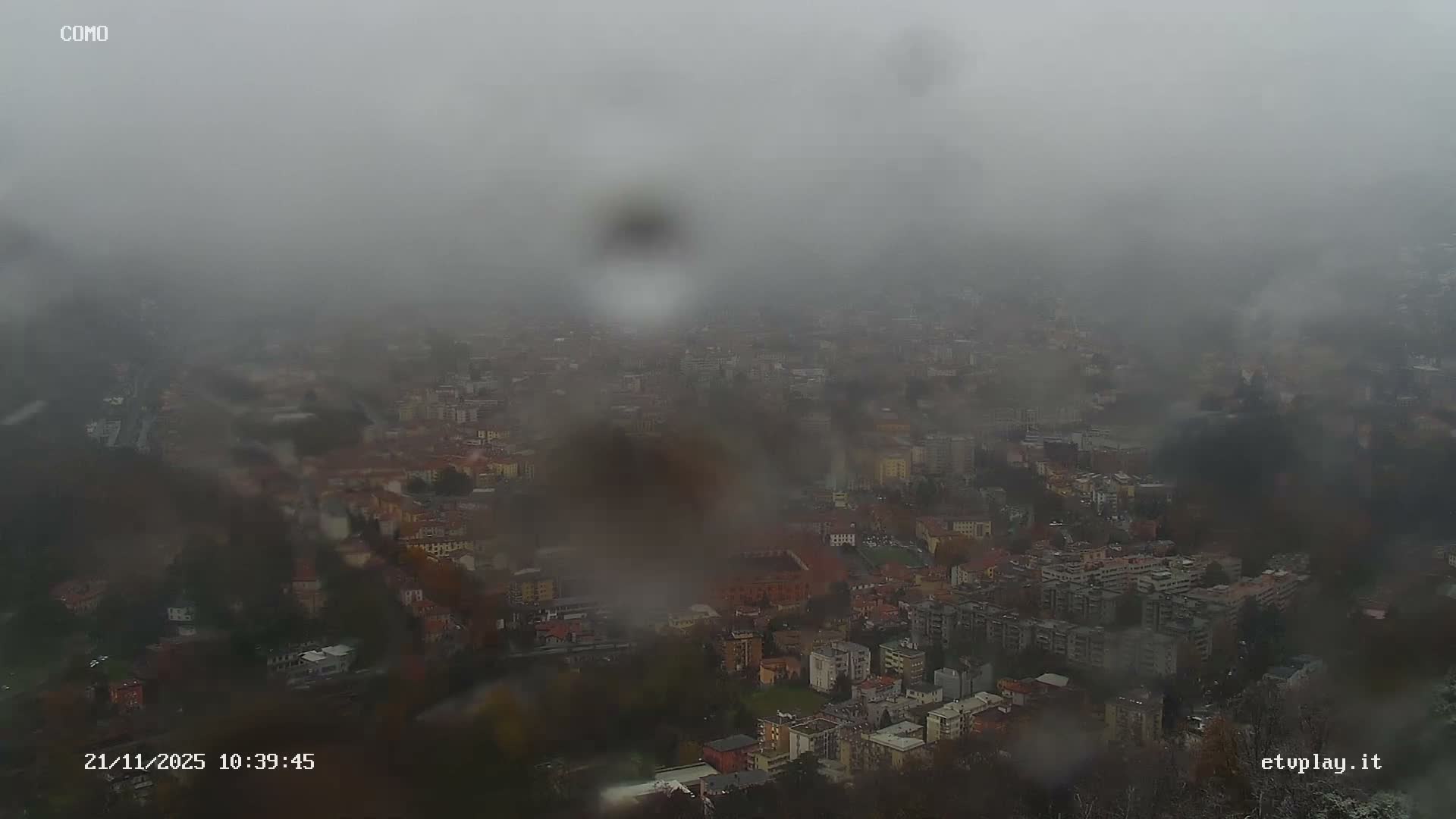 An aerial view of a city is heavily obscured by dense fog and rain droplets on the camera lens, creating a hazy and muted scene.