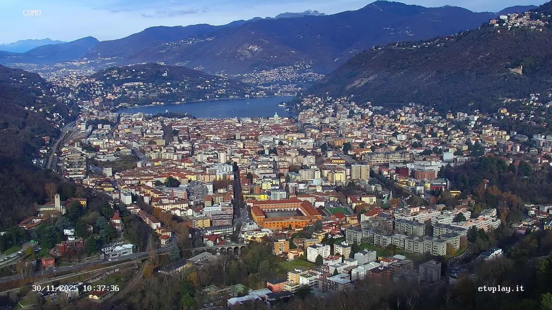 An expansive aerial view captures a densely built city with numerous reddish-brown roofs, nestled between rolling mountains and fronting a large lake under clear skies.