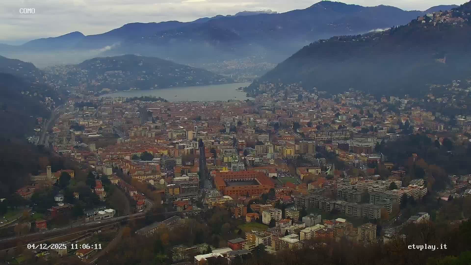 An expansive aerial view reveals a dense city sprawling along the shores of a large lake, encircled by forested mountains shrouded in atmospheric haze under an overcast sky.