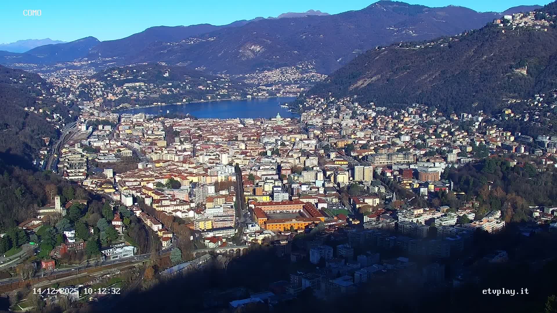 An expansive aerial view reveals a dense city sprawling along the shores of a large lake, encircled by forested mountains shrouded in atmospheric haze under an overcast sky.