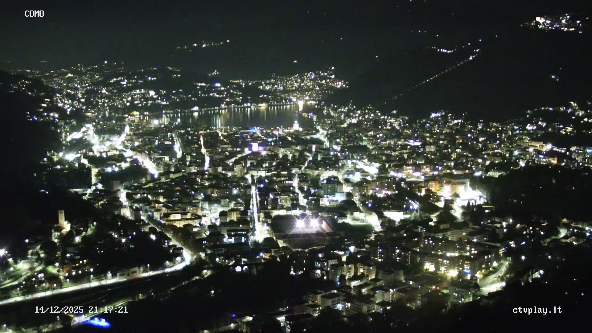 An expansive aerial view reveals a dense city sprawling along the shores of a large lake, encircled by forested mountains shrouded in atmospheric haze under an overcast sky.