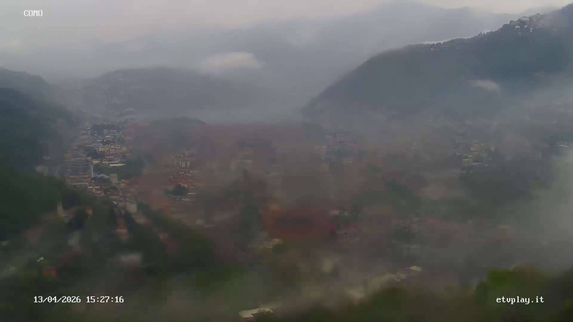An expansive aerial view reveals a dense city sprawling along the shores of a large lake, encircled by forested mountains shrouded in atmospheric haze under an overcast sky.