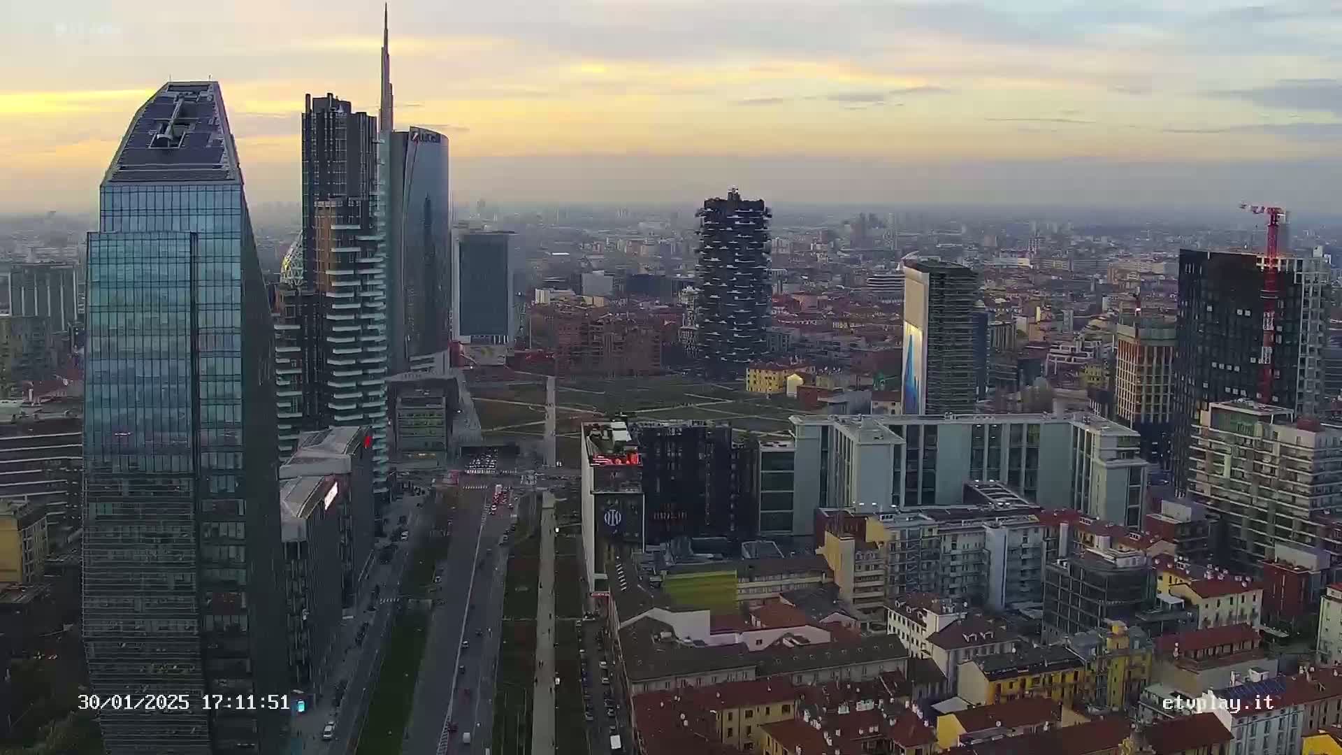 An aerial view of a city skyline at sunset, showing numerous skyscrapers and surrounding buildings under a hazy sky.