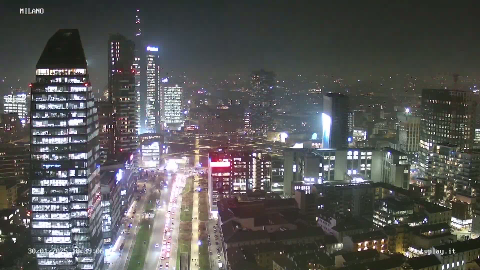 A nighttime aerial view of a city, showing many illuminated skyscrapers and traffic on a wide roadway below, under hazy conditions.