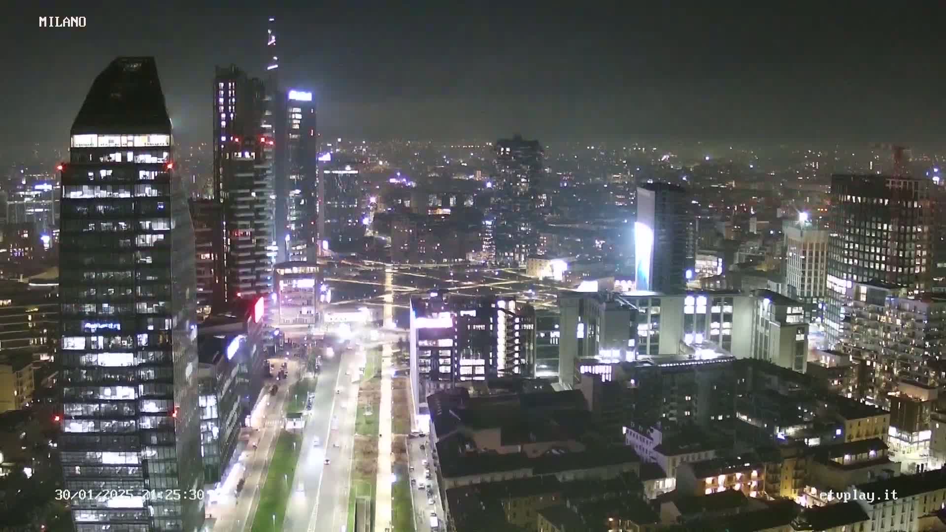 A nighttime aerial view of a city with many skyscrapers and illuminated streets under a hazy sky.