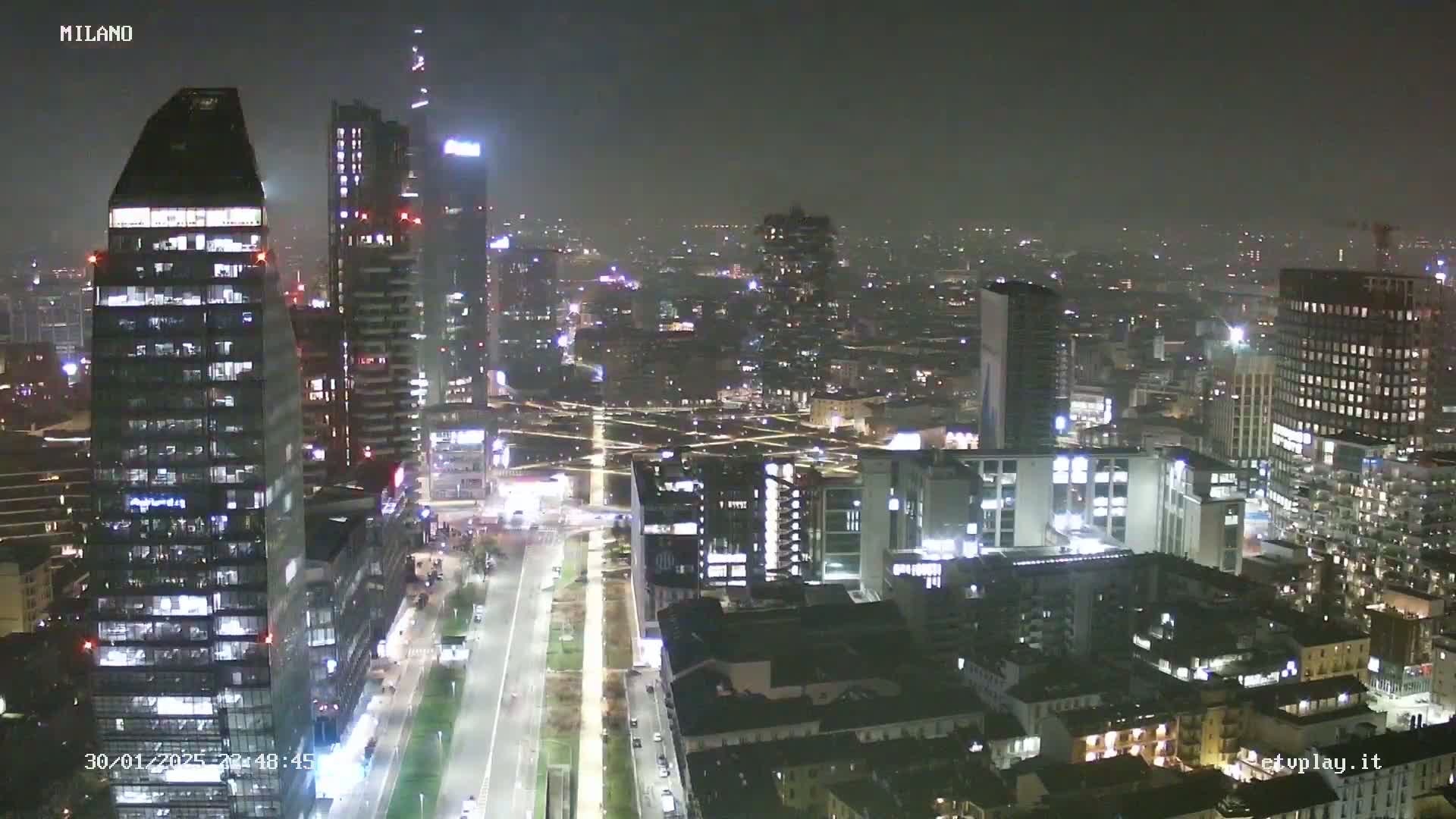 A nighttime aerial view of a city, possibly Milan, shows many illuminated buildings under hazy conditions.