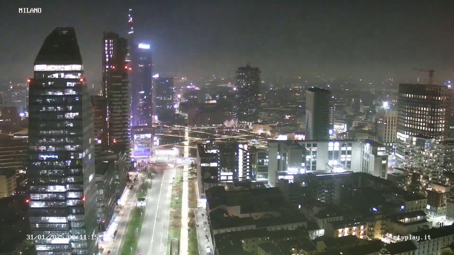 A nighttime, hazy aerial view of a city with numerous illuminated skyscrapers and buildings, and a long, straight road cutting through the urban landscape.