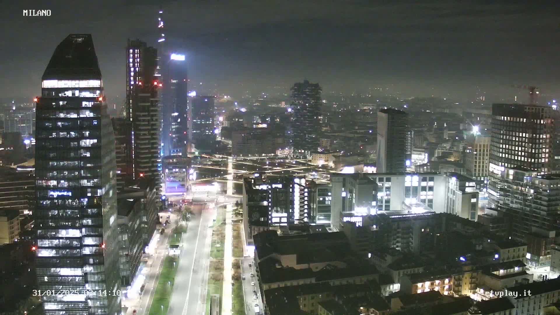 A nighttime aerial view of a city skyline, showing numerous illuminated skyscrapers and buildings under hazy conditions.