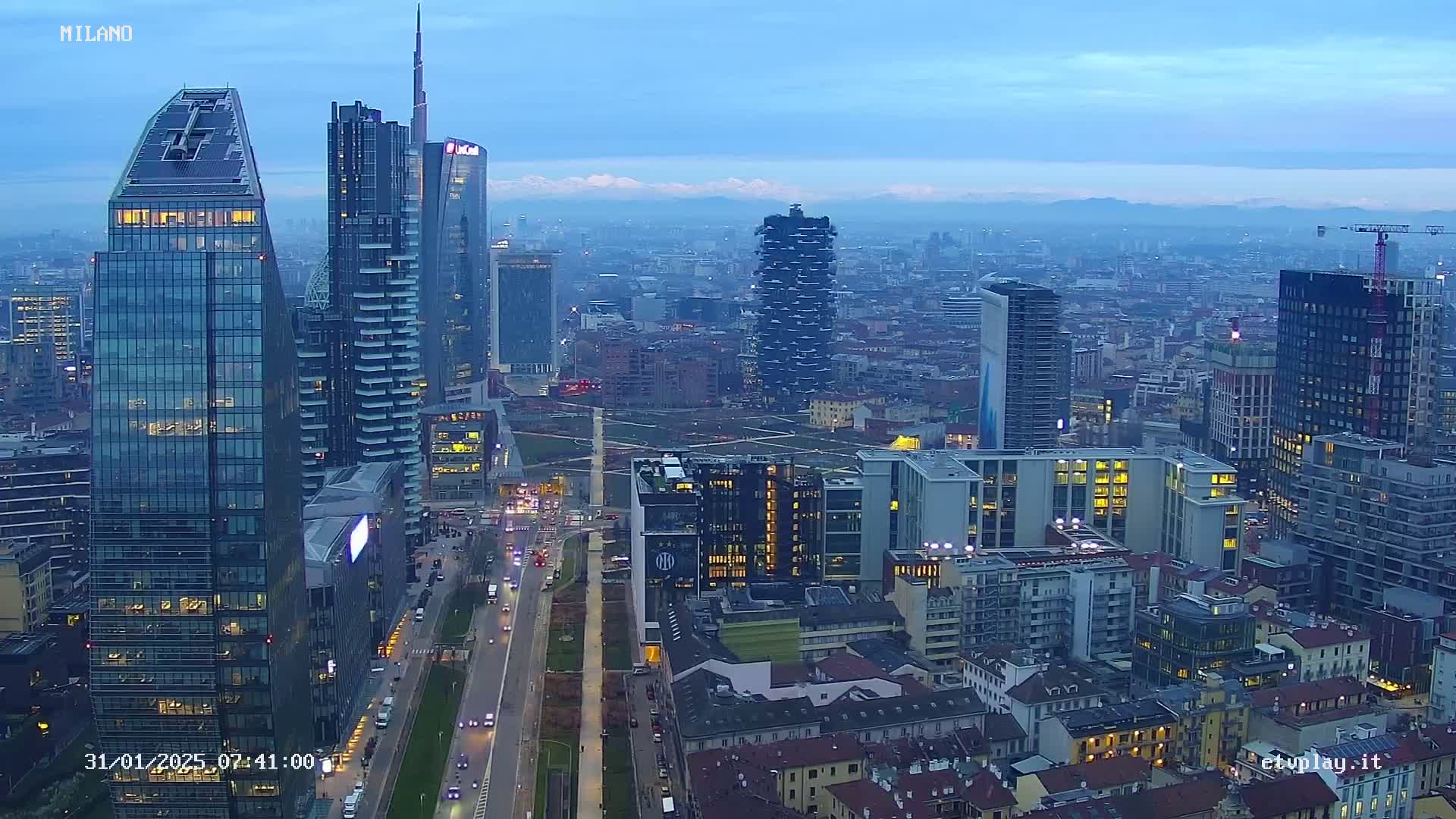 A cityscape at twilight, featuring numerous skyscrapers and city buildings, with mountains faintly visible in the distance under a clear, dusky sky.