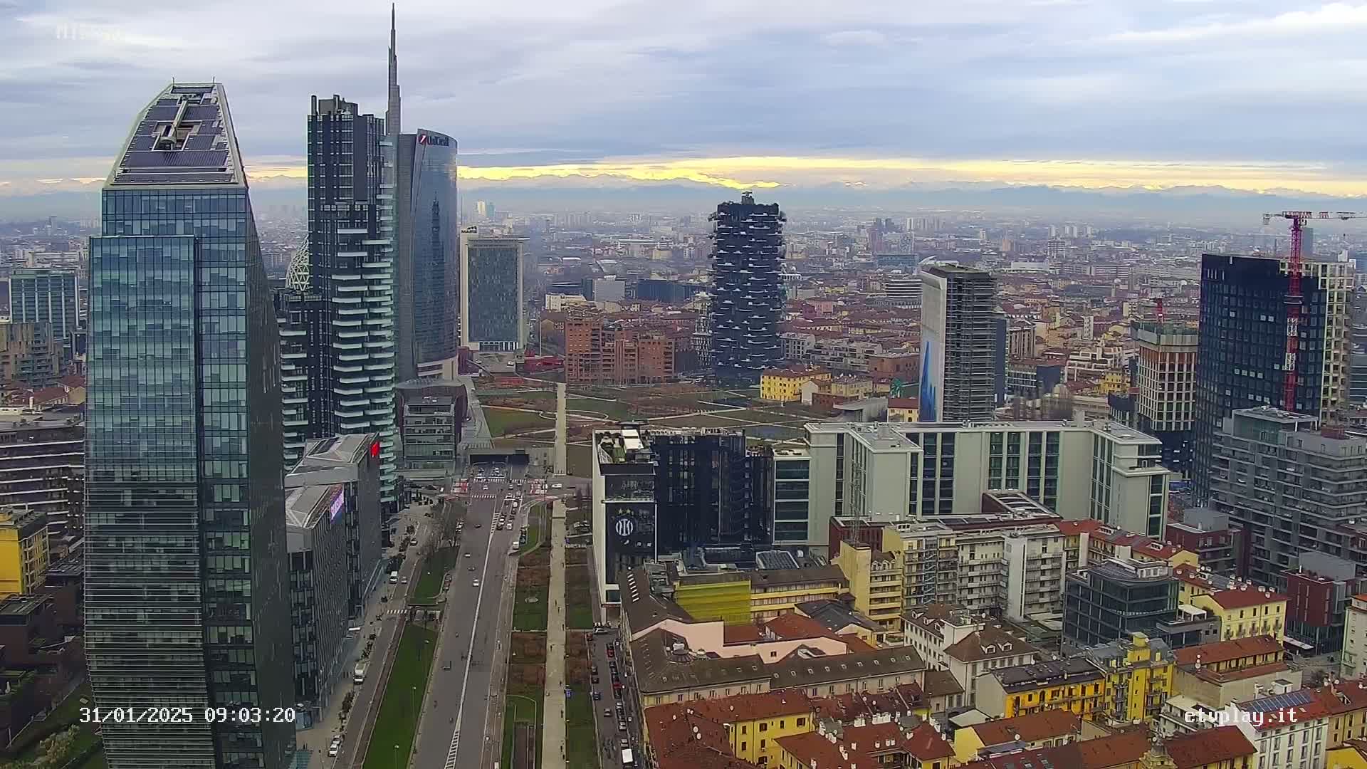 A sprawling cityscape of modern and older buildings under a cloudy sky, with distant mountains visible on the horizon.
