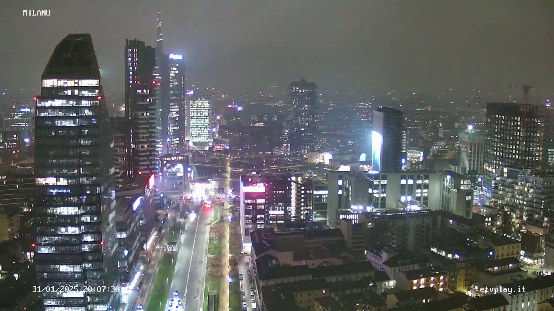A nighttime aerial view of a city skyline with many skyscrapers and illuminated buildings under hazy, overcast conditions.