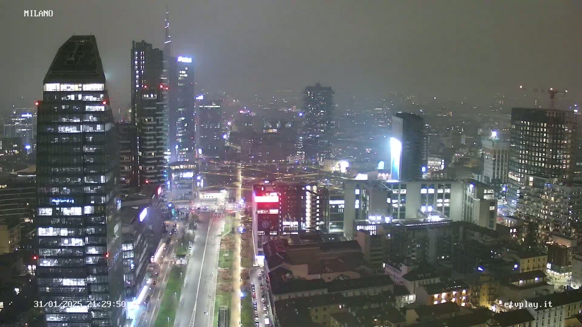 A nighttime aerial view of a city skyline, showing numerous illuminated skyscrapers and buildings under hazy or foggy conditions.