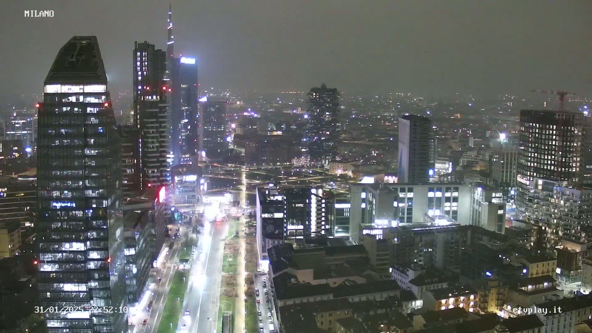 A nighttime aerial view of a city, possibly Milan, showing numerous skyscrapers and city lights under hazy or foggy conditions.