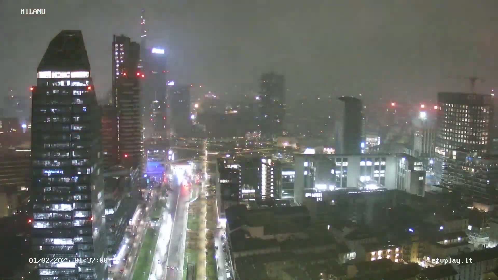 A nighttime, hazy view of a city skyline shows numerous skyscrapers and buildings with lights illuminated, and a wide road below.