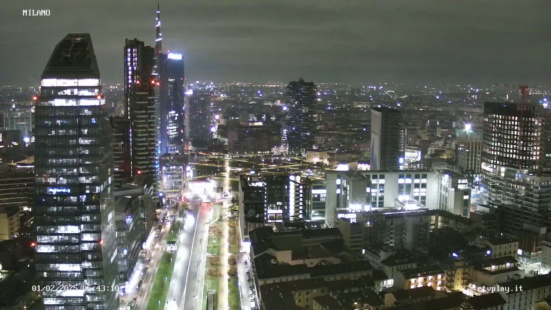 A nighttime aerial view of a city's skyline, featuring numerous illuminated skyscrapers and buildings, under a dark, overcast sky.