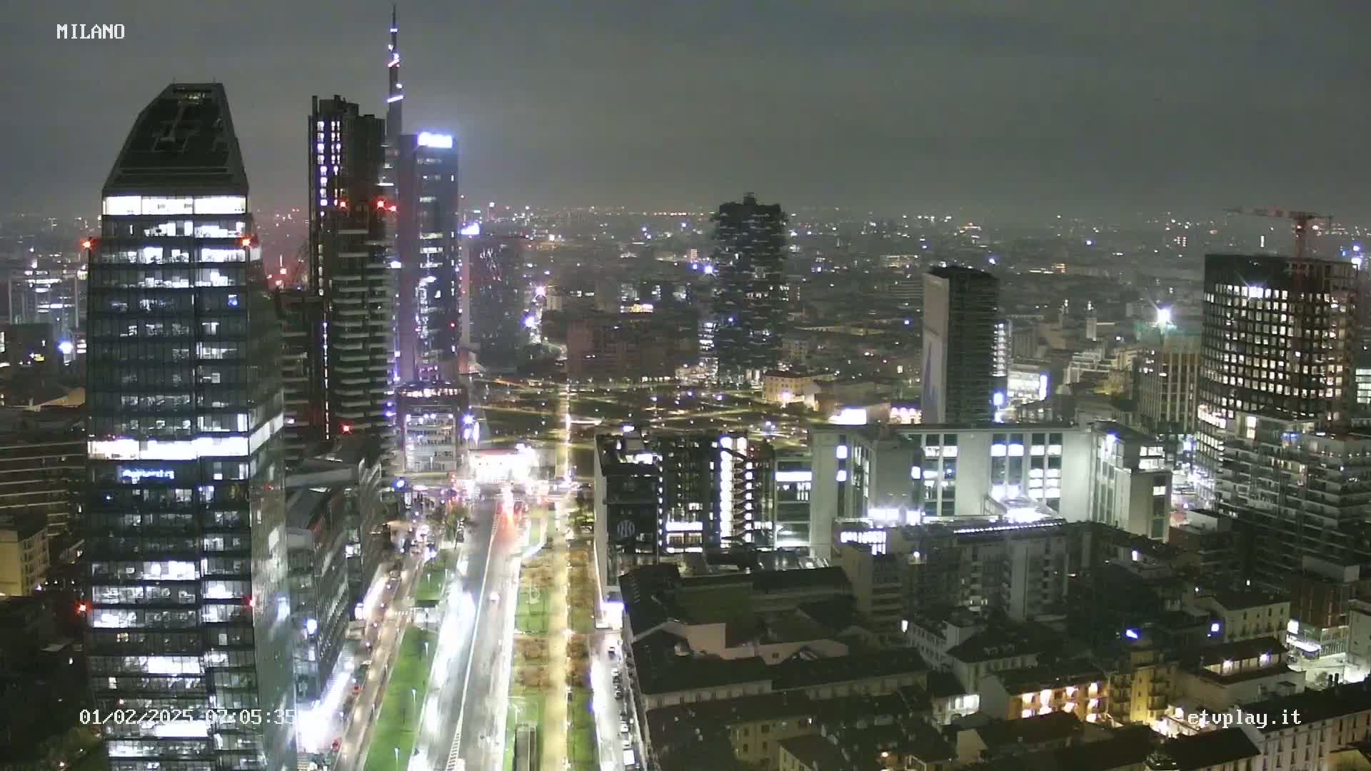 A nighttime view of a city skyline, featuring numerous illuminated skyscrapers and a long, straight road, under hazy conditions.